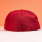 The Big Puma Cardinal White Script New Era Hat 5950 Fitted is shown upside down on a white surface with a peach-colored background, featuring bold Cardinal White Script across the front.