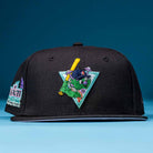 The Flamingo and Chill Bo Bundy 2022 Houston Astros New Era Hat 5950 Fitted is black with a colorful embroidered mascot holding a yellow bat, wearing a helmet, and standing before a teal triangle. It’s shown on a blue surface with a blue background.