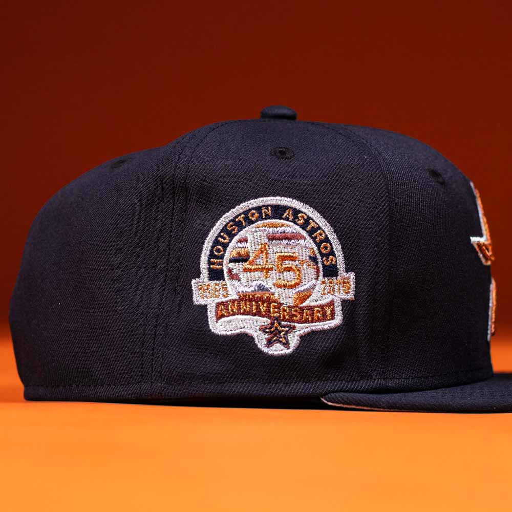 The Highclass 45th Houston Astros New Era Hat 5950 Fitted is a navy blue fitted baseball cap with a 45th anniversary patch on the side, shown on an orange background.
