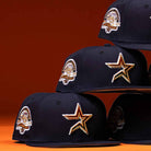 Five Highclass 45th Houston Astros New Era Hat 5950 Fitted caps are stacked in two rows, each black with a gold and white Astros star front logo and “45th anniversary” patch on the side, set against an orange background.