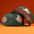 Two green El Jager Mountain Orange New Era 5950 Fitted hats with Texas-themed patches: one has a Houston badge on the side, the other shows a Texas outline with an orange star and Astros logo. Orange background.