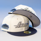 Two International Flea Bo Bundy New Era Houston Astros 5950 Fitted Hats in white and navy, featuring the Astros logo on the front and an “Astrodome The Original” patch on the side, stacked against a light blue background.