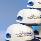 Four white International Flea Bo Bundy Houston Astros New Era 5950 Fitted hats with blue brims are stacked. Each features "Astros" embroidered on the front and a side patch reading "Astrodome The Original" against a light blue background.