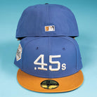 Two Harbor Mark Canvas Houston Astros New Era 5950 Fitted Hats are stacked. The front one shows ".45s" in white with a side patch, and the back displays the MLB logo. Both have blue crowns, brown brims, and sit on a light blue background.