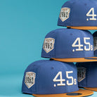 A stack of Harbor Mark Canvas Houston Astros New Era Hat 5950 Fitted caps with gold brims, featuring ".45s" on the front and a Houston 1962 patch on the side, displayed against a blue background.