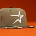The Pre-Order Wall Hanger New Era Bark Real Tree Houston Astros 5950 Fitted Hat features an olive green body, white star front logo, and camouflage brim, shown on an orange background. Now available for pre-order with easy shipping options.