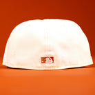 The Rustic Sunset Houston Astros New Era Hat 5950 Fitted is shown upside down, highlighting the small MLB logo on the back, displayed against an orange background.