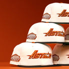 A stack of Rustic Sunset Houston Astros New Era Hat 5950 Fitted caps featuring orange and gold Astros logos and Astrodome The Original patches is displayed on an orange background.