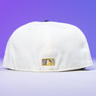 The Trilltiberi Rushmore WS22 Chrome Graphite Houston Astros New Era Hat 5950 Fitted is shown from the back with a gold and black MLB logo patch at the bottom center, set against a purple gradient background. Classic meets modern in this standout cap.
