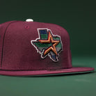 The Pinkerton 2.0 Rushmore 2000 Maroon Houston Astros New Era Hat 5950 Fitted features an embroidered Texas outline with a multicolored, stylized star on the front, inspired by Astros colors and set against a dark green background.