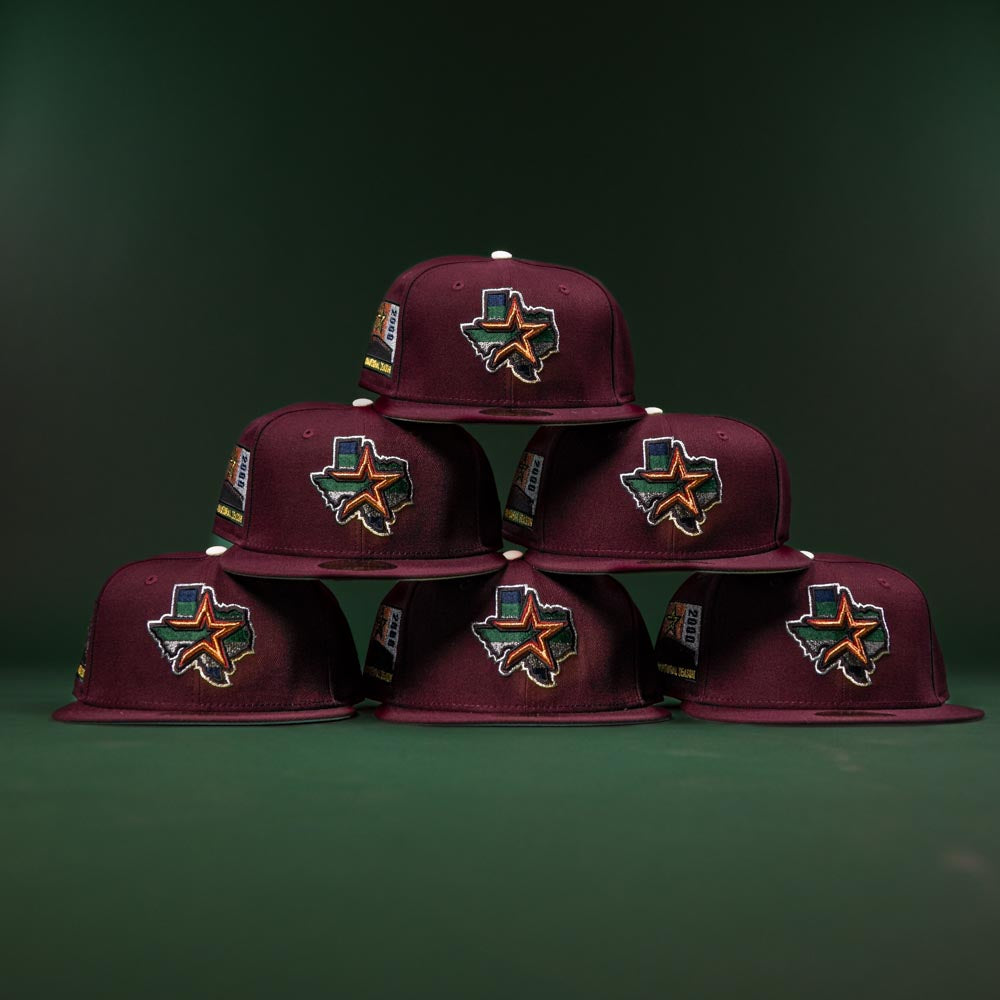 Six maroon caps, including the Pinkerton 2.0 Rushmore 2000 Maroon Houston Astros New Era Hat 5950 Fitted, are stacked in a pyramid. Each features vibrant Texas patches, embroidered details, and flag accents on a dark green background.
