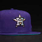 The DAY N HOU Rushmore WS05 Purple Teal Houston Astros New Era 5950 Fitted Hat features a large embroidered H with a gold star on the front, a flat brim with teal underside, and a bold black and purple design.