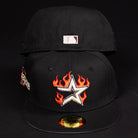 Two black La Flame Rushmore WS05 Fire Black Houston Astros New Era Hat 5950 Fitted caps on a black background—one displays a silver star with red-orange flames, and the other has a small white and brown logo on the back.