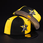 Two Hazmat Rushmore Gold Black Pinwheel Houston Astros 5950 Fitted New Era Hats are shown: one upright and one tilted to display the underbrim, both featuring a stylized H logo, set against a plain black background.