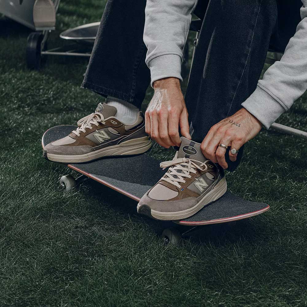 A person sitting outdoors ties the laces of 933 GRY Andrew Reynolds Gray Day New Balance Numeric suede skateboarding shoes while resting their feet on a skateboard placed on grass.