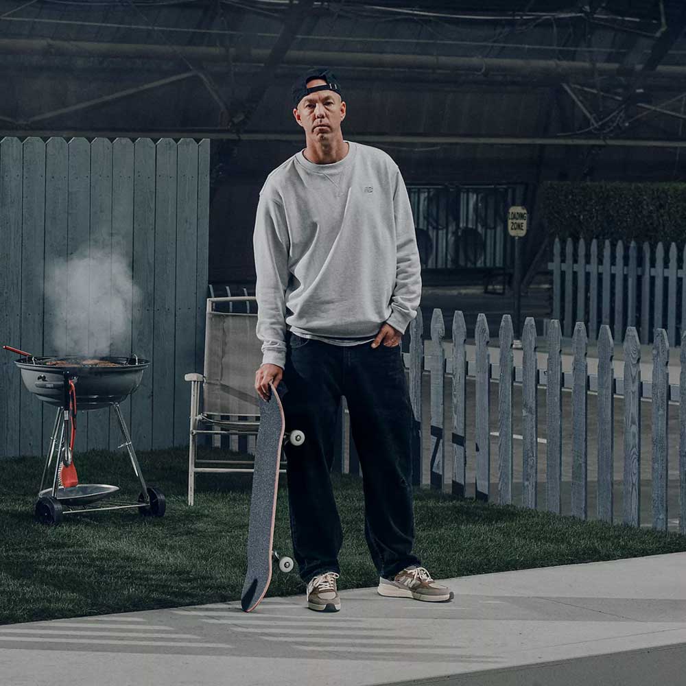 A man in a backward cap, gray sweatshirt, and dark pants stands outdoors holding a skateboard in his 933 GRY Andrew Reynolds Gray Day New Balance Numeric Suede Skateboarding Shoes beside a smoking grill, with lawn chairs and a picket fence in the background.