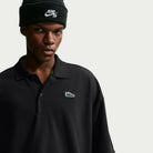 A person wearing a black Nike SB AF1 Black Court Green Solid Break Polo IV0597010 with a small sneaker emblem on the chest and a black beanie.