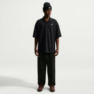 A person wears a loose-fitting black AF1 Black Court Green Solid Break Polo IV0597010 Polo Top Nike SB Skateboarding, black trousers, a black beanie, and dark sneakers.