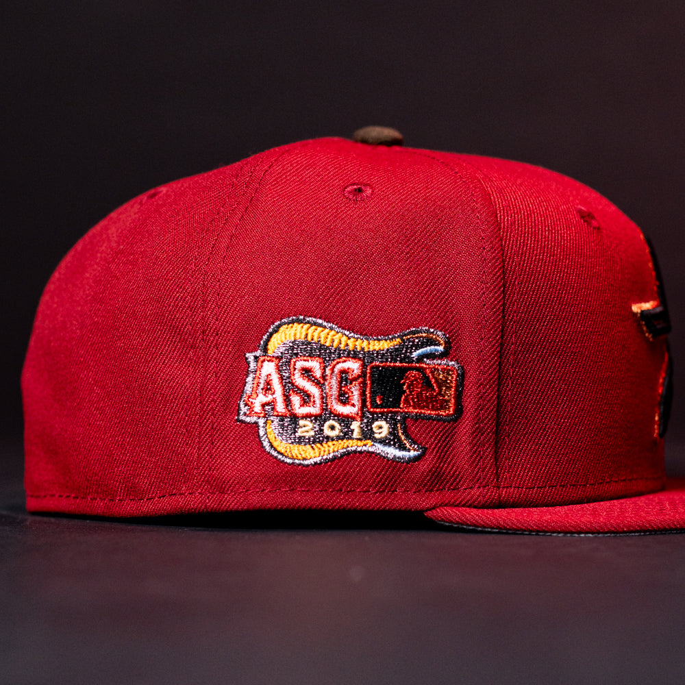 The ASG19 Bred Houston Astros New Era Hat 5950 Fitted is a red cap with a guitar-shaped ASG 2019 patch and an MLB logo embroidered on the side, celebrating the Astros. The dark, blurred background highlights its details.