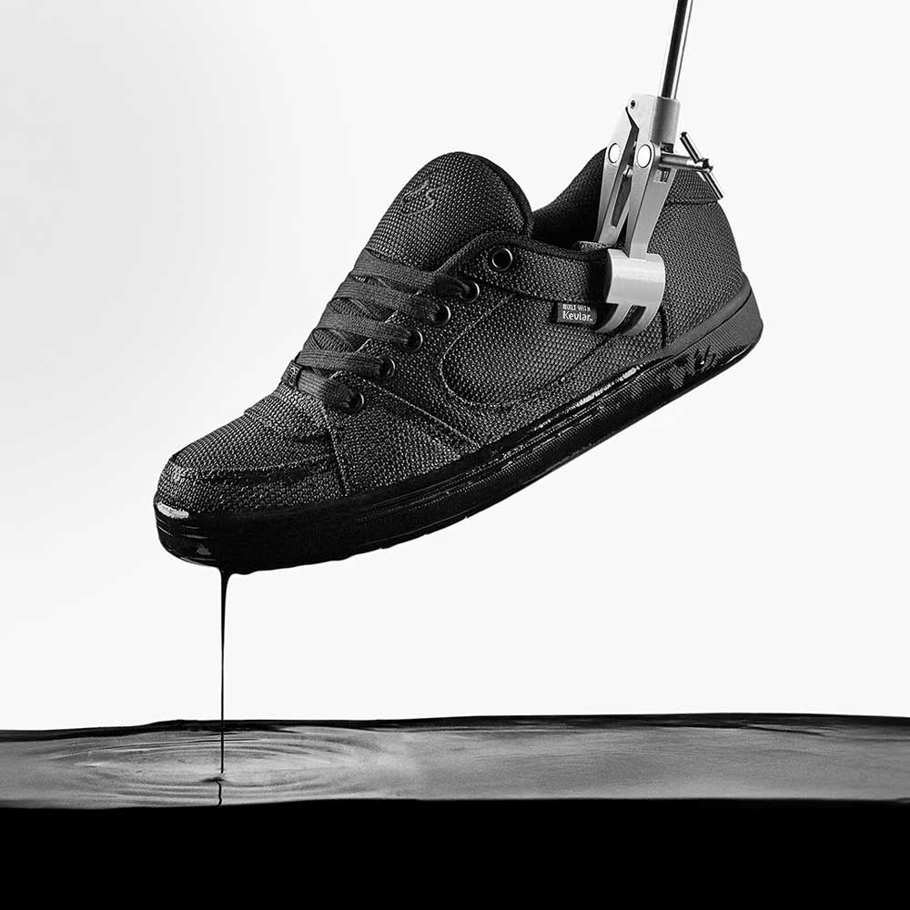 A black Accel Kevlar Black Es Skateboarding Shoe is held by a metal clamp above a pool of black liquid, with drops falling from the shoe and making ripples on a minimal white background.