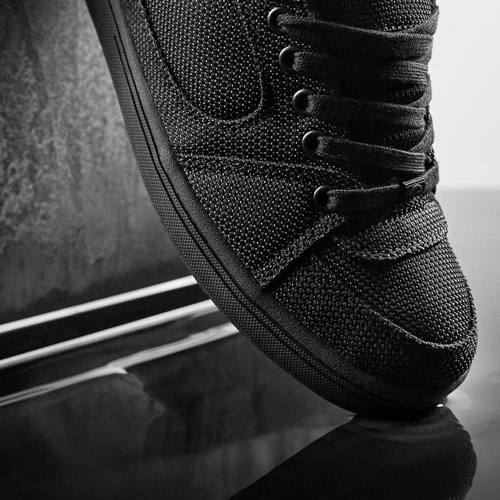 Close-up of the Accel Kevlar Black Es Shoes Skateboarding, featuring a black textured design with thick laces, shown side view at an angle and reflected on a glossy black surface with a metal background.