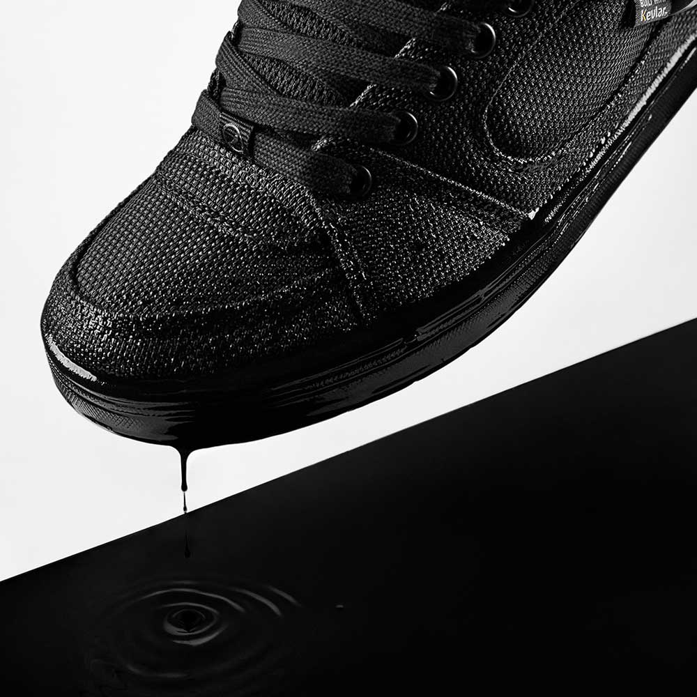 A close-up of Accel Kevlar Black Es Shoes Skateboarding, with water droplets beading on the surface and dripping into a reflective black puddle below, creating a ripple effect.