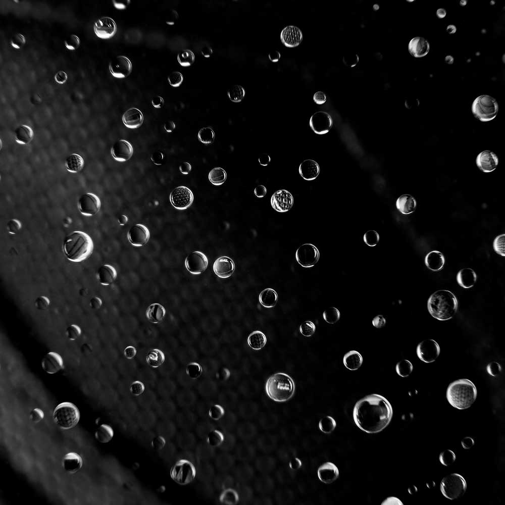 Close-up of water droplets scattered on a dark surface, each reflecting light and subtle patterns reminiscent of the Accel Kevlar Black Es Shoes Skateboarding, creating a monochrome, abstract impression.