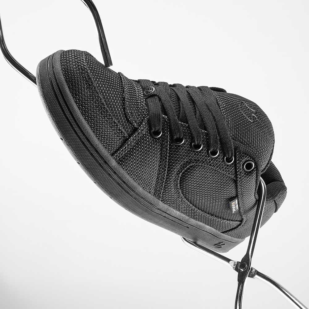 The Accel Kevlar Black Es Skateboarding Shoes, featuring textured black fabric and laces, are balanced on two metal rods against a white background, showcasing timeless skate style.