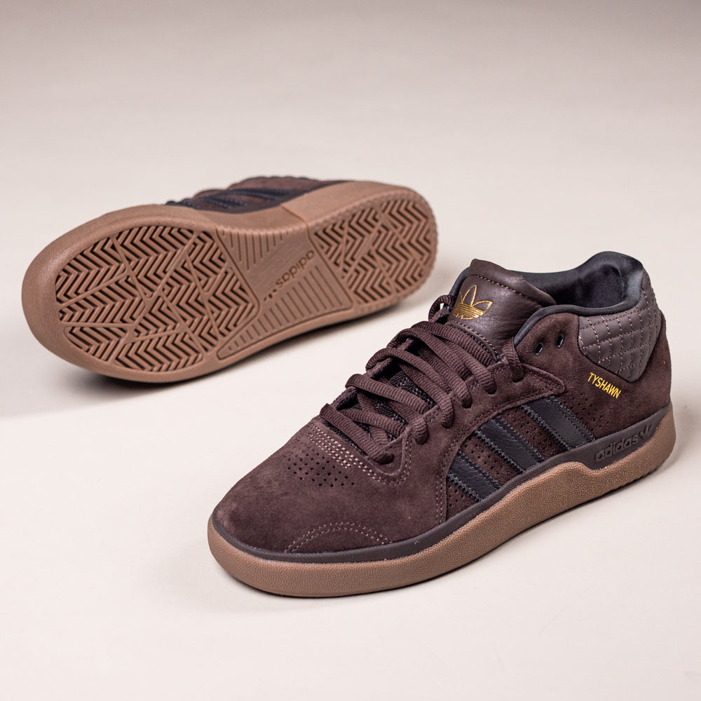 The Tyshawn HQ4740 Brown Gum Adidas Skateboarding Suede Shoes, featuring black stripes, gold branding, and gum rubber soles, are shown on a neutral background with one upright and the other on its side.