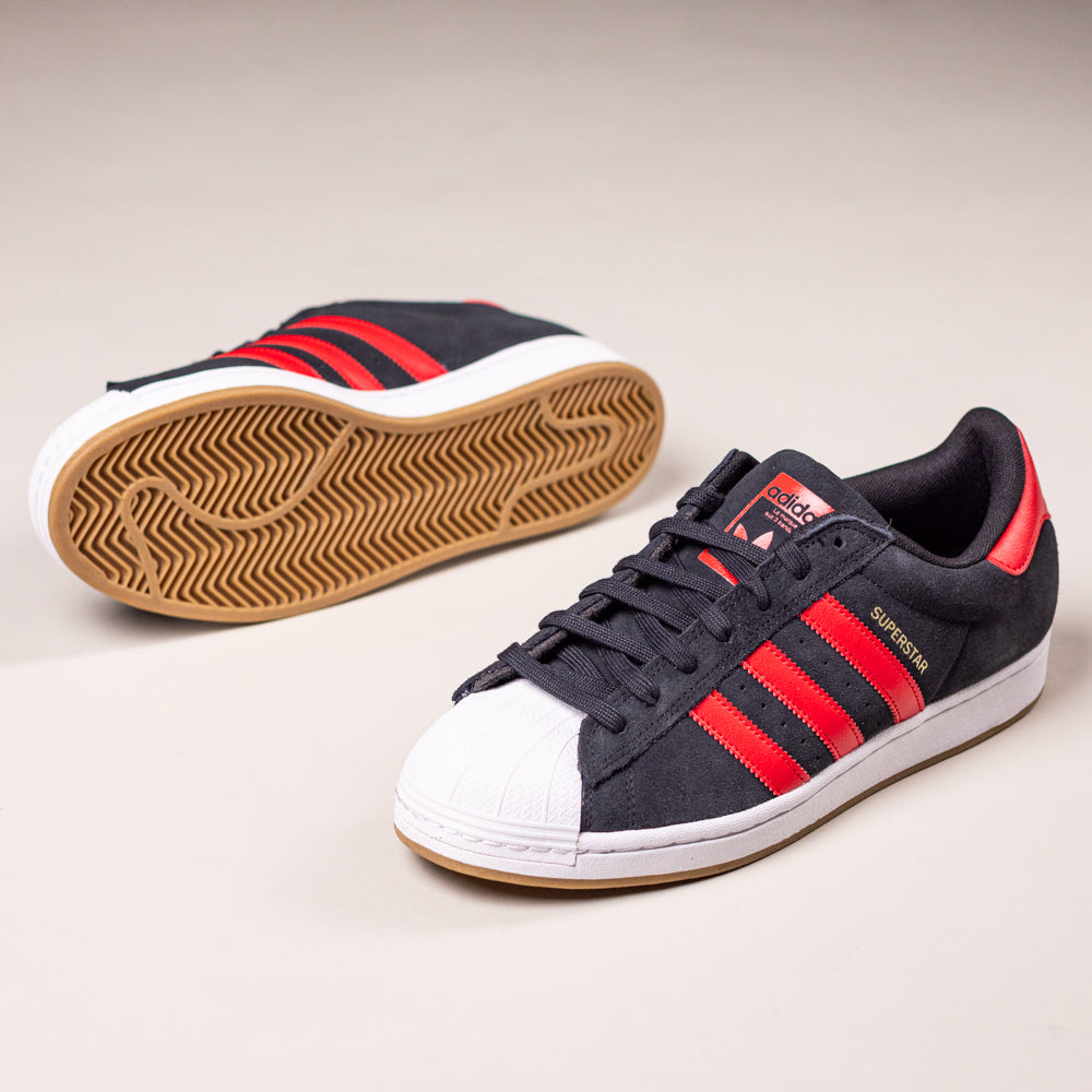 A pair of Adidas Superstar ADV Black Red KI5731 suede leather skateboarding shoes with black uppers, red stripes, white rubber toe caps, and tan outsoles—one upright, one on its side showing the sole.
