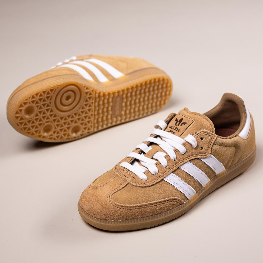The KI5089 Tan White Samba ADV Adidas Skateboarding Suede Shoes are displayed on a beige surface, one upright and one on its side to highlight the textured gum rubber sole, ideal for skateboarding enthusiasts.