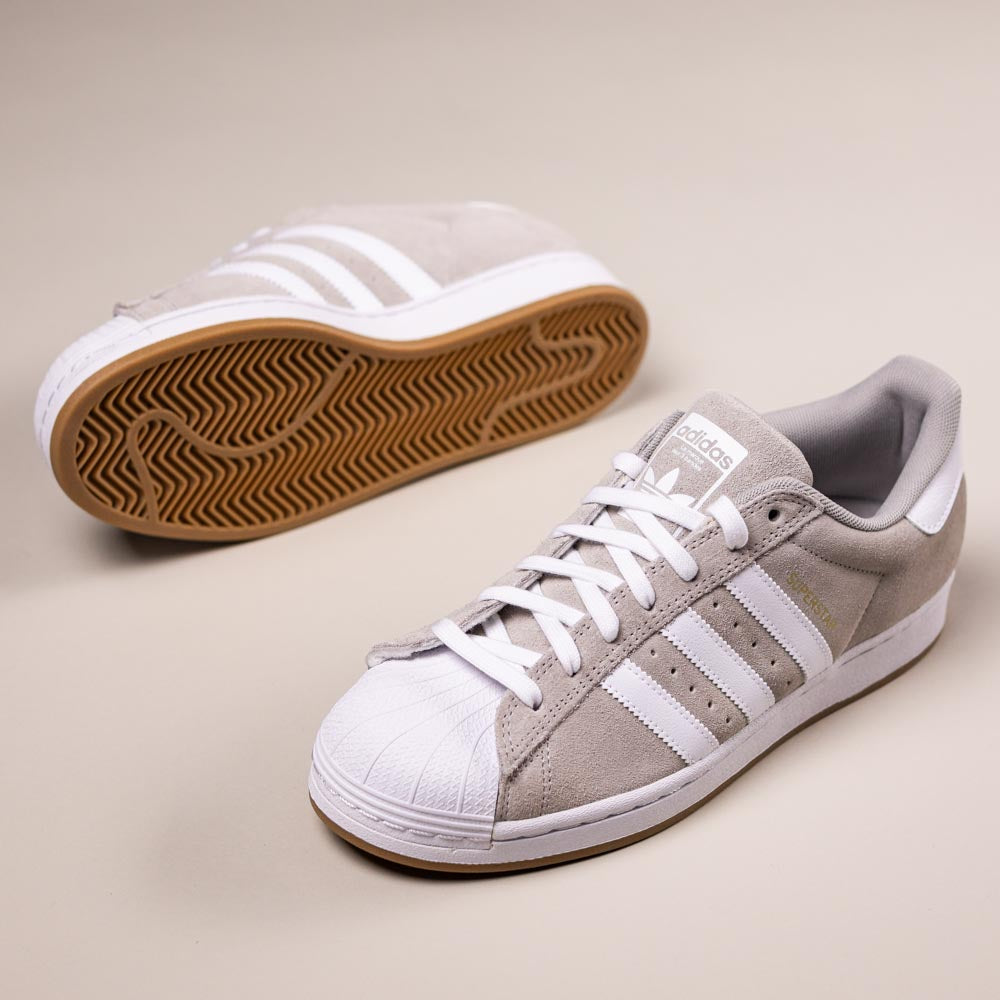 The KI5733 Gray White Superstar ADV Adidas Skateboarding Suede Shoes feature light gray suede uppers, white stripes and laces, and gum rubber soles. They are displayed on a beige background, one upright and one lying on its side.