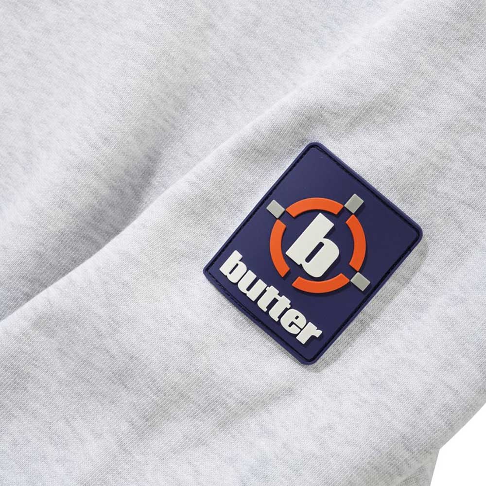 A close-up of the Aleka Ash Crewneck Butter Goods Sweatshirt, a grey cotton outerwear featuring a rectangular patch with a blue background, orange and white target design, a white lowercase "b," and "butter" below in white letters.