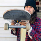 A person in a black beanie and plaid shirt holds a skateboard towards the camera, showcasing the Andrew Allen Hollow Silver Black Standard INDY 144 Stage 11 Trucks Set of TWO, with their face out of focus in the background.