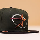 The GWOP Dark Green Burgundy Bronze Circle Star Houston Astros New Era Hat 5950 Day features a dark green crown, black brim, and an embroidered orange star outlined in gold with a white swoosh on the front. The background is soft beige.