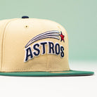 The New Era Hat 5950 Houston Astros Vegas Gold Emerald Aeros 5950 Day features ASTROS in blue, a red star, white shooting star streaks on the front, an Astrodome sidepatch, a tan crown, and a green flat brim with a light green background.