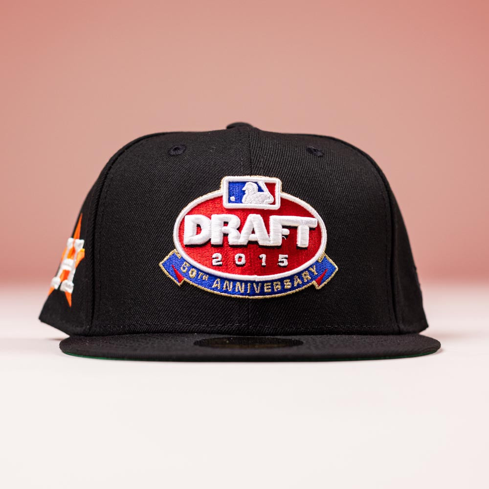 A black New Era 5950 Fitted Hat featuring the Houston Astros 2015 Draft Day design in red and blue, shown against a pink background.