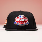 A black New Era 5950 Fitted Hat featuring the Houston Astros 2015 Draft Day design in red and blue, shown against a pink background.