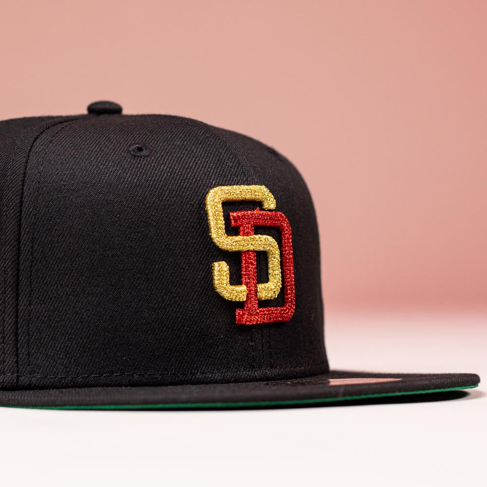 The Bones Pack San Diego Padres New Era 5950 Fitted Hat features a black design with the iconic yellow and red SD logo embroidered on the front, displayed against a soft pink background.