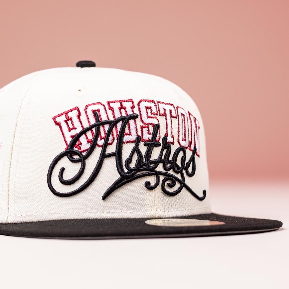 The D BOY Houston Astros New Era 5950 Fitted Hat in ivory, black, and red is shown against a pink background.
