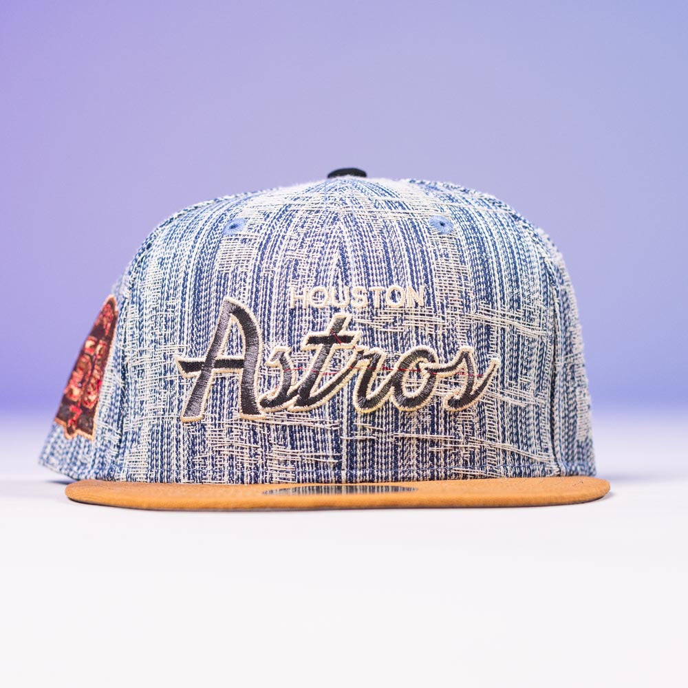 A Bronze Denim 50th Houston Astros Hat New Era 5950 Fitted, featuring Astros embroidery and a tan brim, is shown against a light purple background.