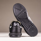 A pair of Leggerezza Asics Black Bright White 1201B045 leather skateboarding shoes; one stands upright to reveal the heel, while the other leans forward, highlighting the tread pattern and durable leather upper.