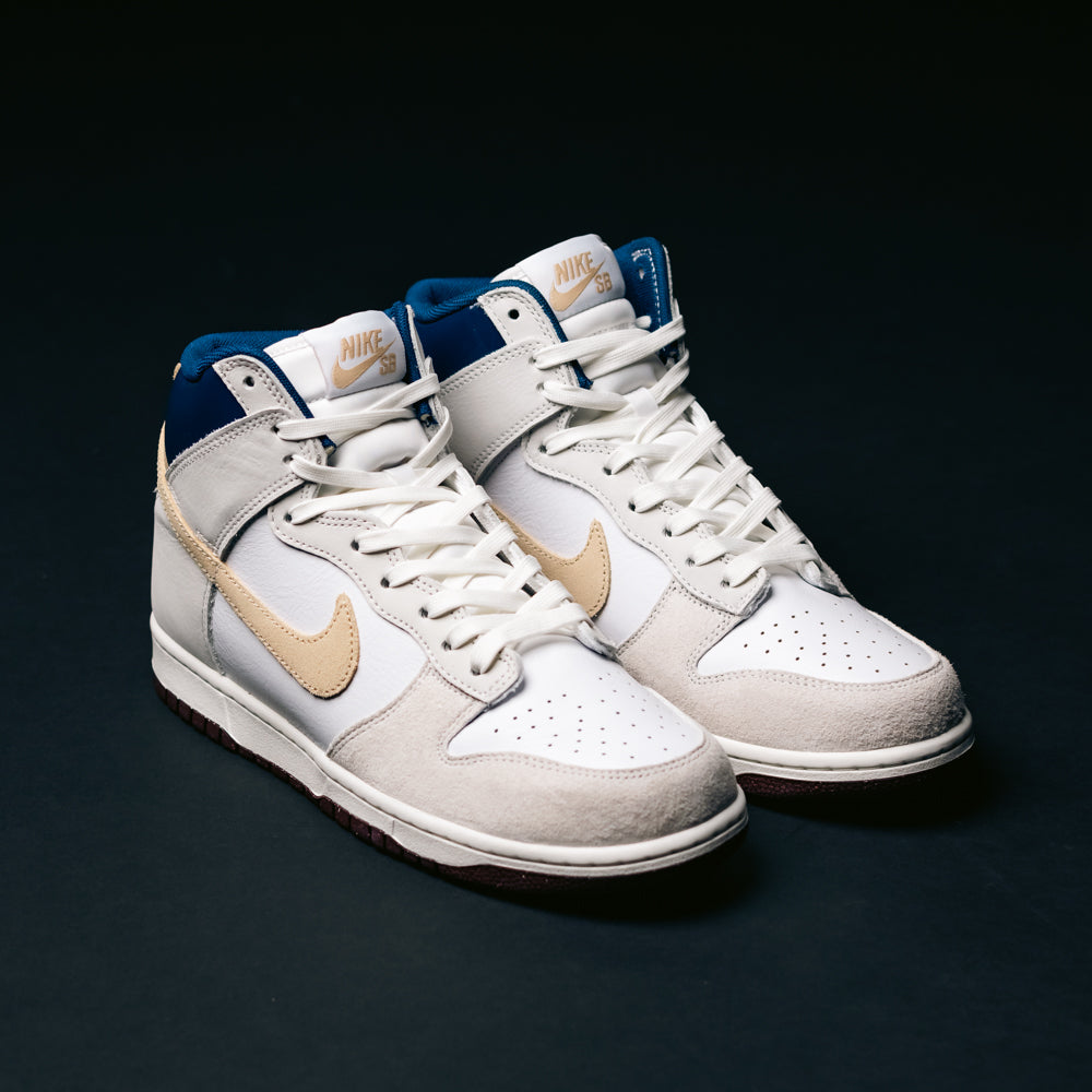 A pair of Ivory Sand Blue Void Dunk High Pro x Beyond IU4491100 Nike SB Skateboarding Shoes, featuring white leather, light gray suede, tan swooshes, and navy blue ankle details, are placed on a dark surface.