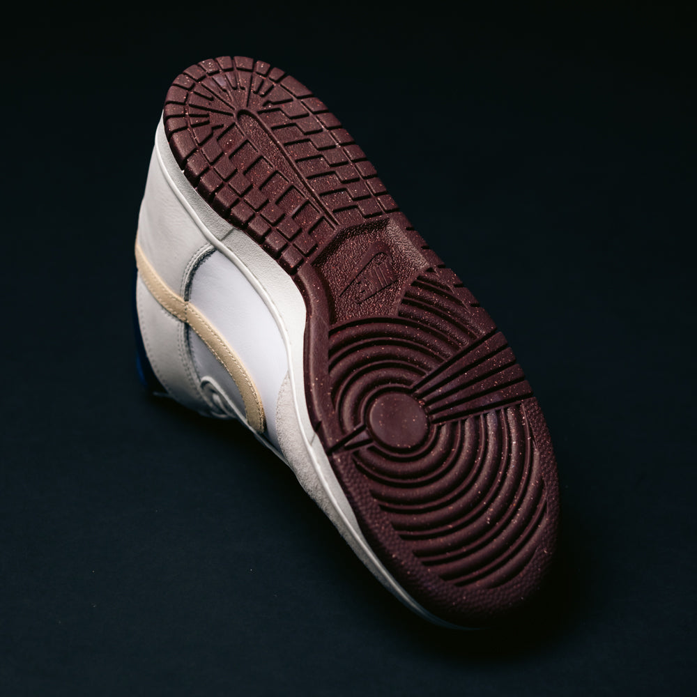 The Ivory Sand Blue Void Dunk High Pro x Beyond IU4491100 Nike SB Skateboarding Shoe is displayed with its burgundy sole facing upward, showcasing the distinctive tread pattern against a dark background.