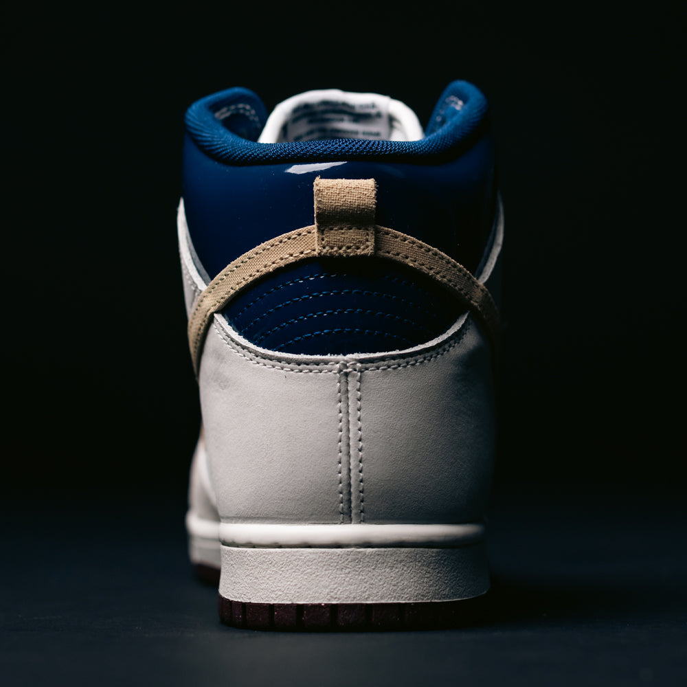 A close-up of the back of the Nike SB Dunk High Pro x Beyond IU4491-100 "Ivory Sand Blue Void" skateboarding shoe, showcasing its white, beige, and dark blue colorway with a beige pull tab and dark sole, set against a dark background.