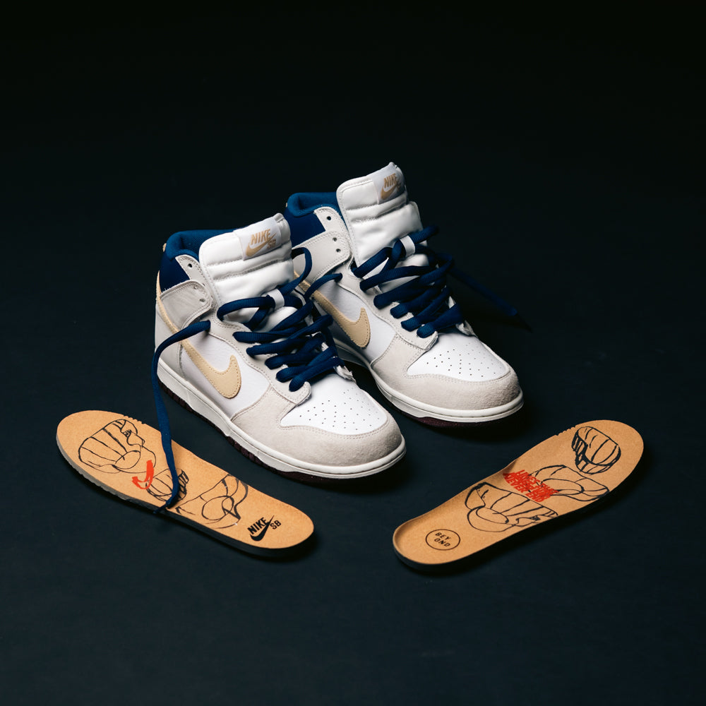A pair of Ivory Sand Blue Void Dunk High Pro x Beyond IU4491100 Nike SB skateboarding shoes are displayed with navy laces beside insoles that showcase cartoon-style artwork, all set against a black background.
