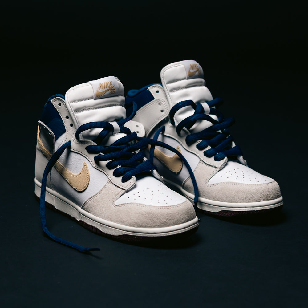 A pair of Nike SB Dunk High Pro x Beyond IU4491100 "Ivory Sand Blue Void" skateboarding shoes, featuring white, beige, and navy blue panels with gold swooshes, dark blue laces, and padded white tongues, placed on a dark surface.