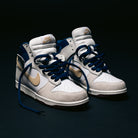 A pair of Nike SB Dunk High Pro x Beyond IU4491100 "Ivory Sand Blue Void" skateboarding shoes, featuring white, beige, and navy blue panels with gold swooshes, dark blue laces, and padded white tongues, placed on a dark surface.