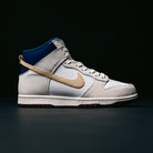The Ivory Sand Blue Void Dunk High Pro x Beyond IU4491100 Nike SB Skateboarding Shoes feature a stylish basketball-inspired design with white and gray panels, a tan swoosh, blue accents on the collar, and a dark sole, displayed against a dark background.