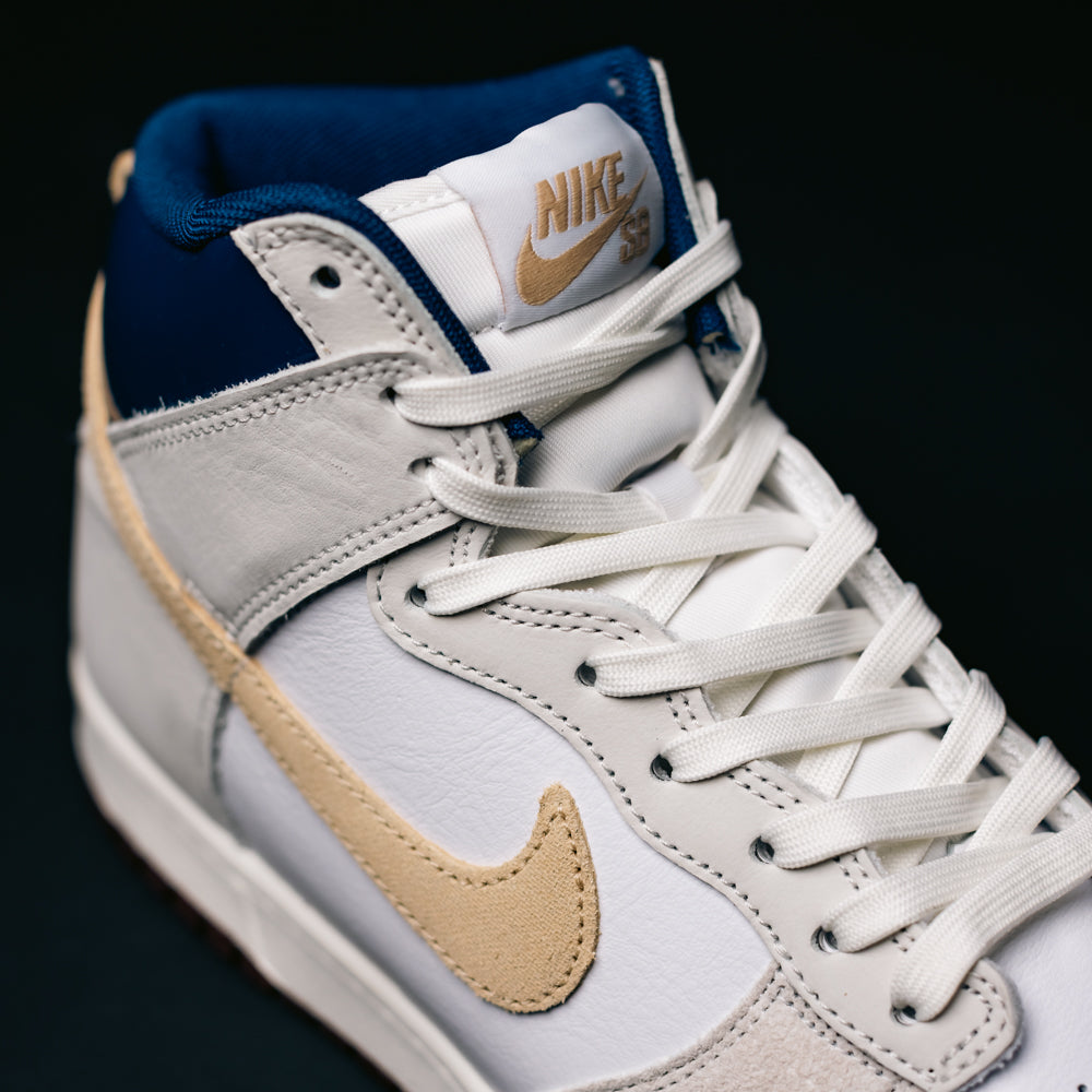 Close-up of the Ivory Sand Blue Void Dunk High Pro x Beyond IU4491100 Nike SB Skateboarding Shoe, featuring a white and beige colorway with blue accents, white laces, a beige Nike Swoosh, and gold Nike branding on the tongue against a dark background.
